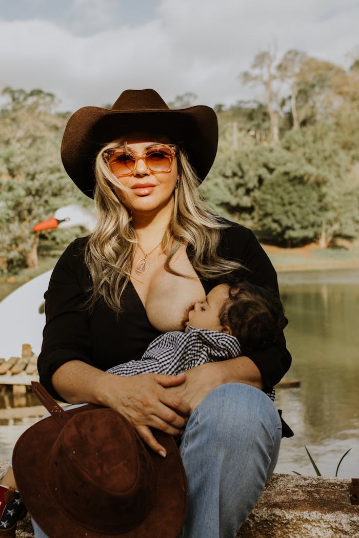 A mother in cowboy attire breastfeeding her baby by a serene lake outdoors.