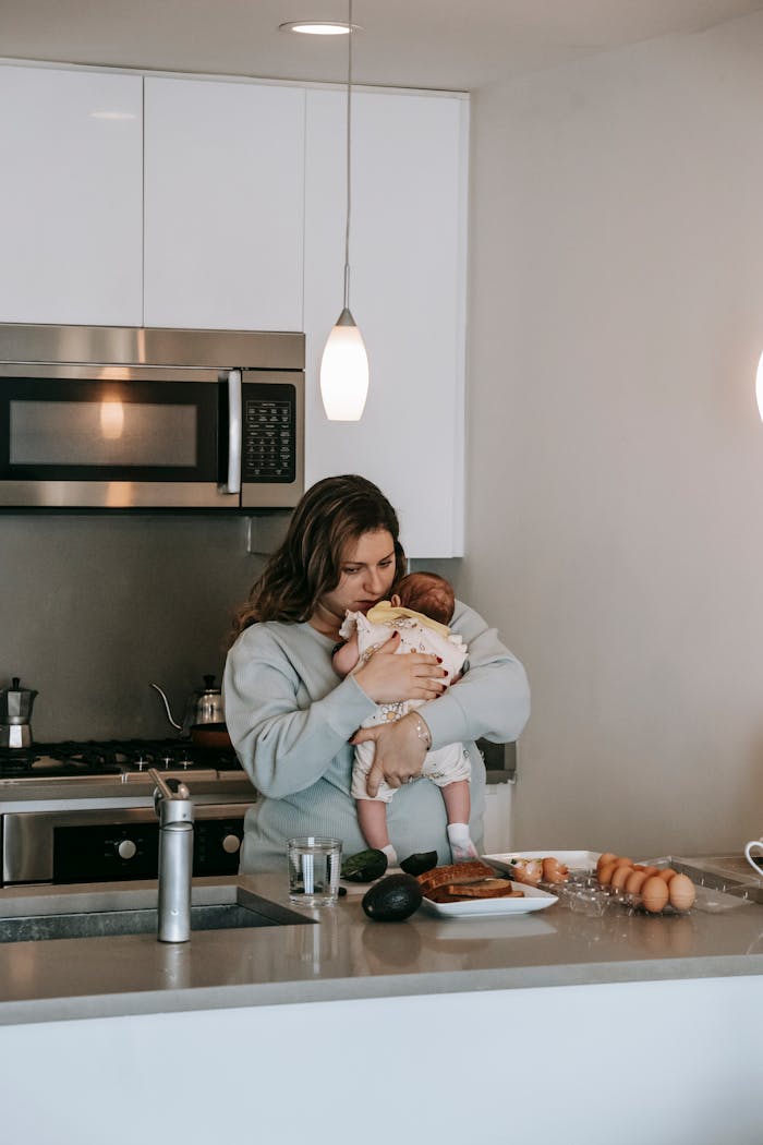 Loving young mother with long hair in casual clothe hugging cute baby while standing in kitchen during breakfast preparation
